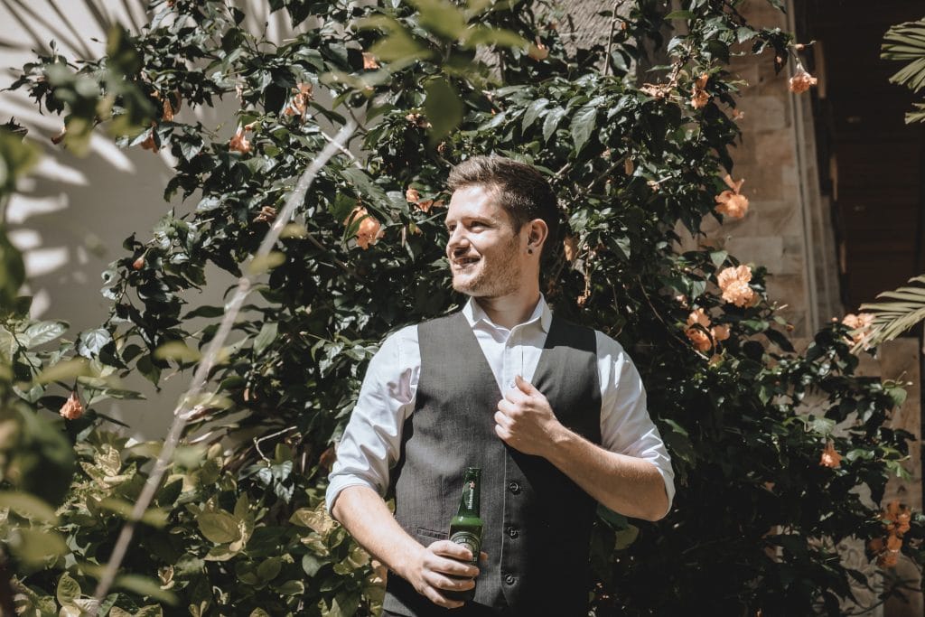 A man holding a bottle of beer in front of bushes.