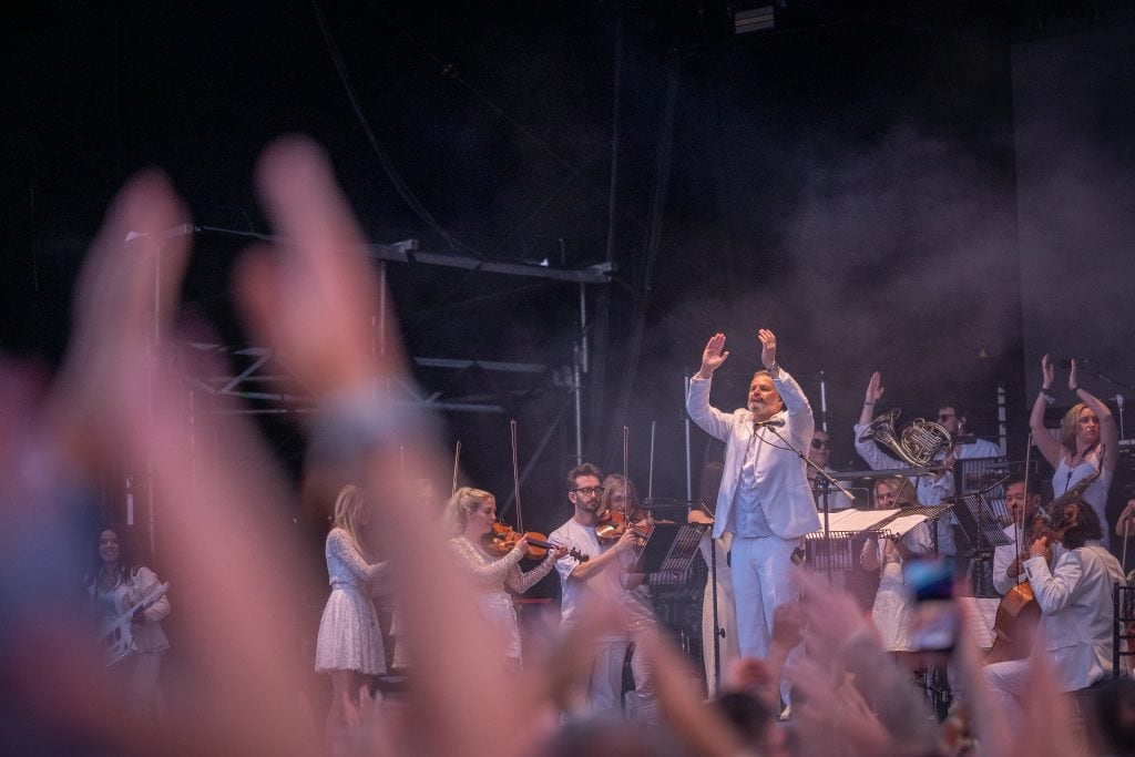 A man in a white suit with his hands up in the air at a concert.