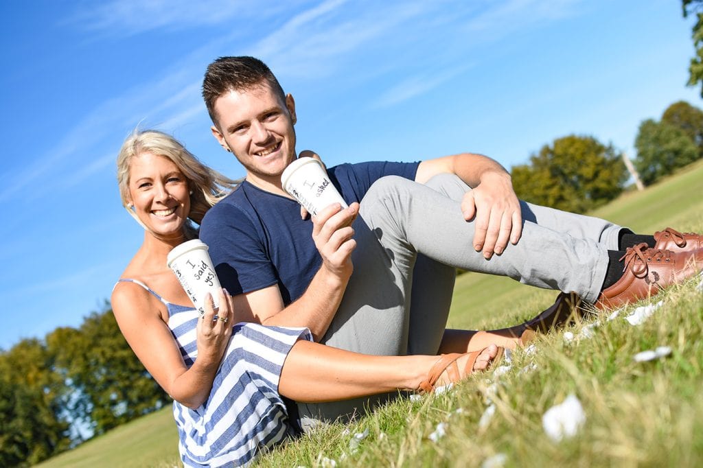 A man and woman sitting on a grassy field holding a cup of coffee.