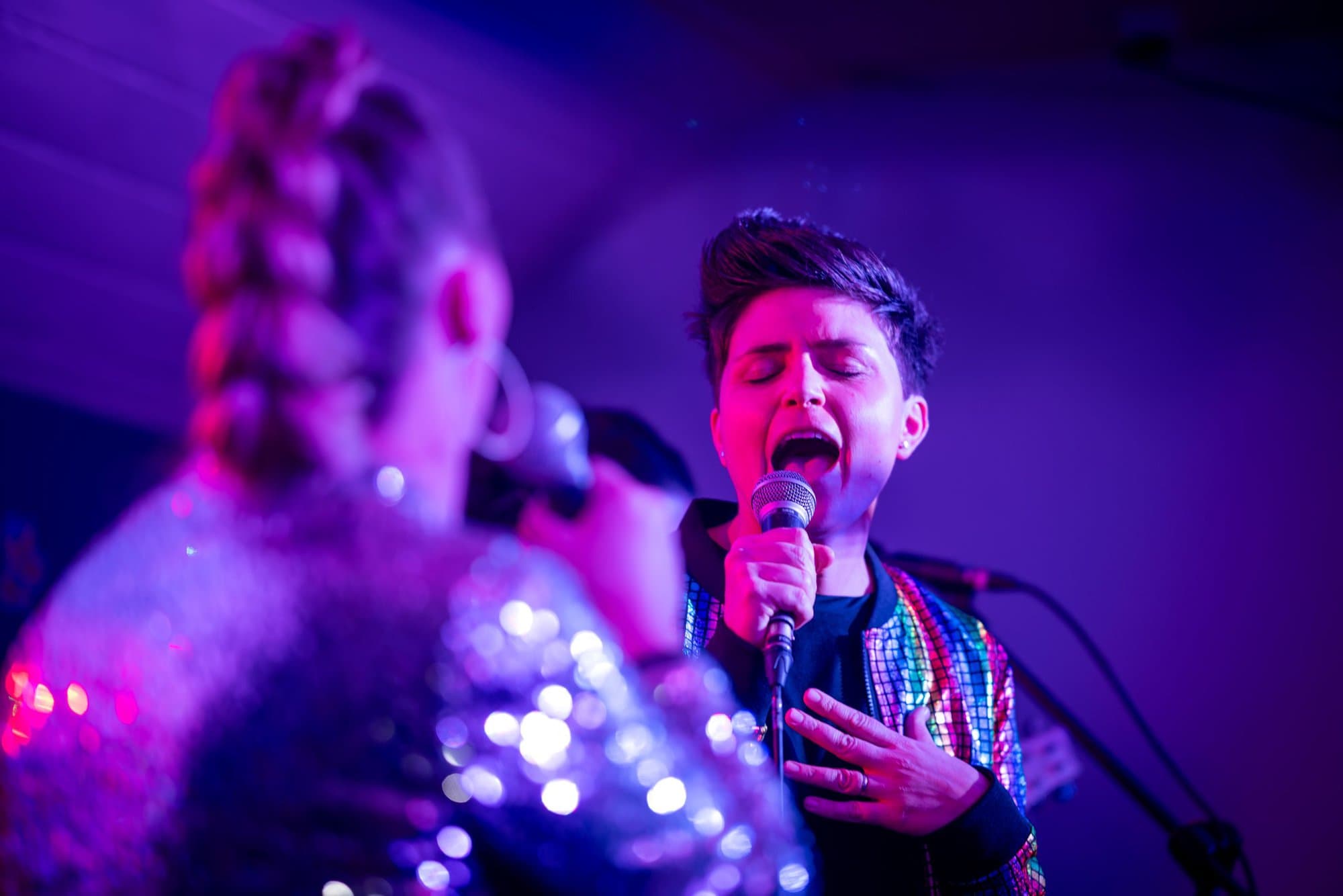 Two singers performing onstage with one holding a microphone passionately singing into it under purple stage lighting.