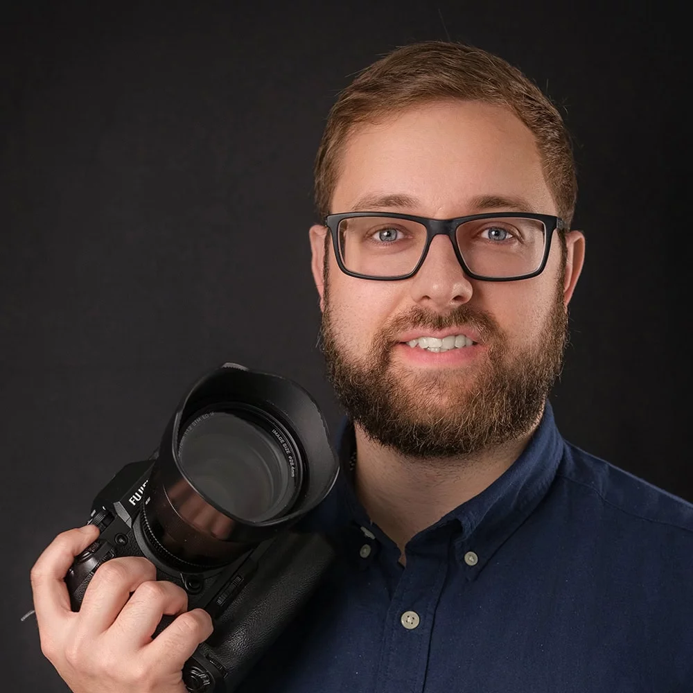 Man with glasses and beard, wearing a blue shirt, holds a professional camera against a dark background.