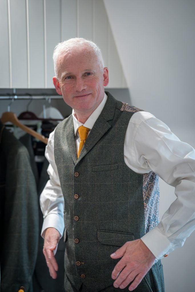 A man in a patterned waistcoat and tie stands indoors, adjusting his outfit. A hanging jacket is seen in the background.