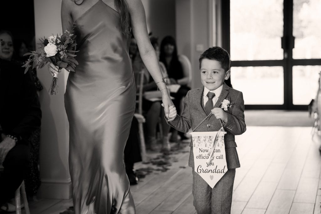 A young boy in a suit holds a sign reading "Now you can officially call me your Grandad" while walking hand-in-hand with a woman in a dress at an indoor event.