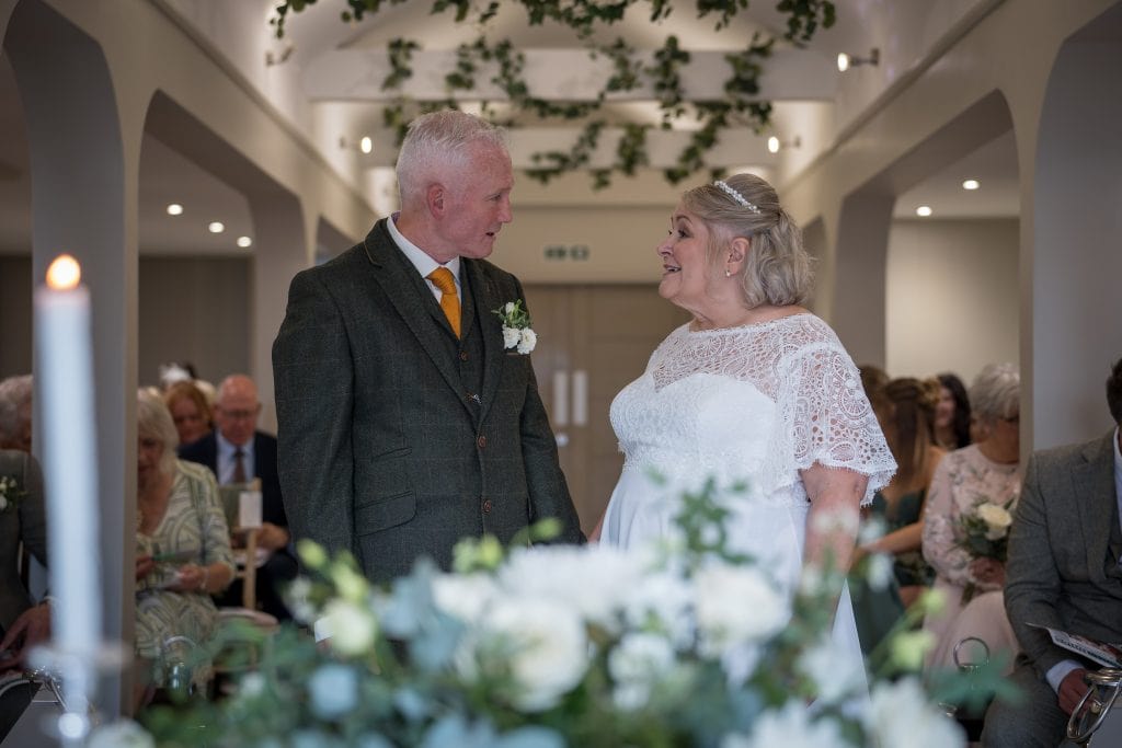 An elderly couple in wedding attire stands facing each other in a decorated indoor ceremony with guests seated in the background.