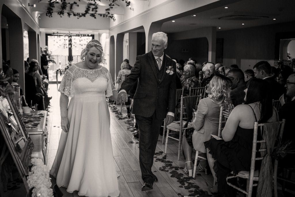 A bride and groom walk down the aisle hand in hand at a wedding ceremony, surrounded by seated guests in a decorated indoor venue.