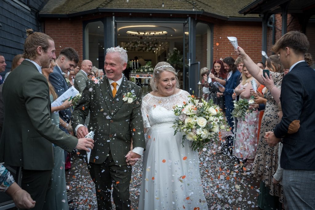 A couple walks hand in hand through a crowd of people throwing confetti, outside a building. The bride holds a bouquet, and the groom is smiling.