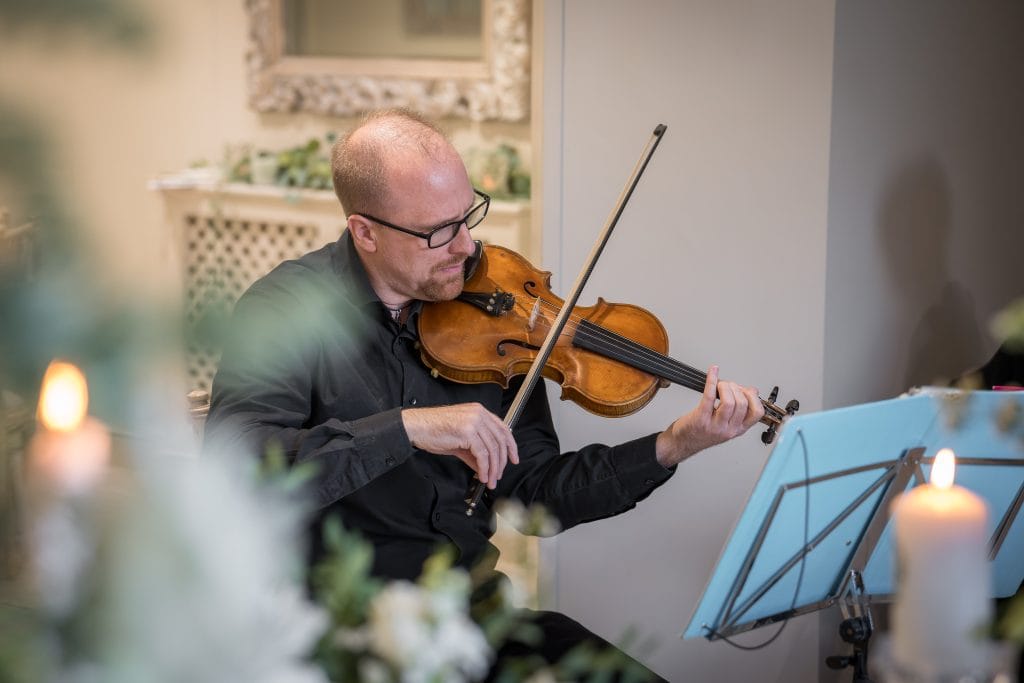 A man wearing glasses plays a violin while reading sheet music, with blurred candles and greenery in the foreground.