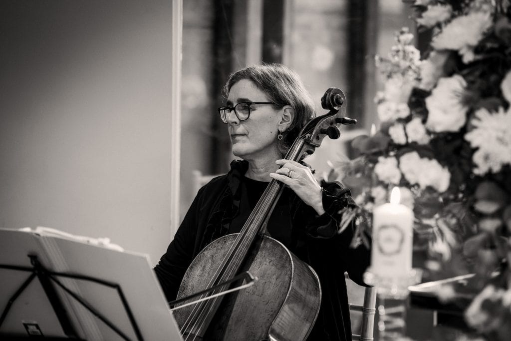 Cellist wearing glasses plays the cello, seated beside a stand with sheet music. Flowers and a lit candle are visible nearby.