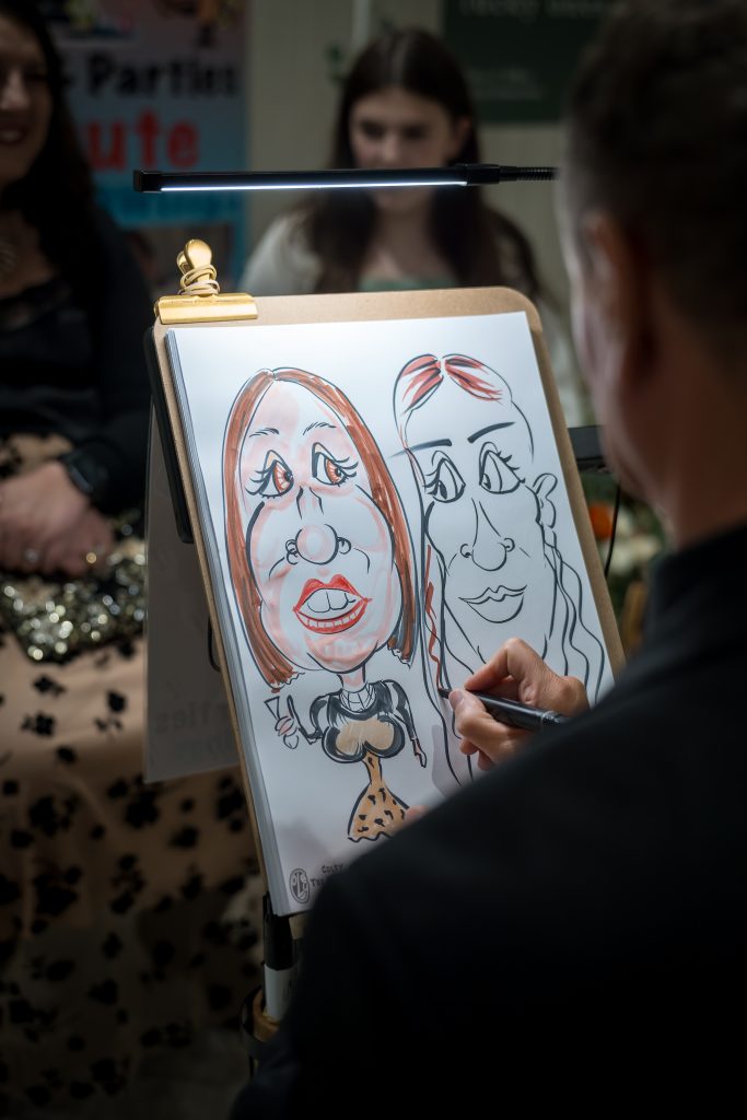 An artist sketches two caricatures of women on a drawing board as three people watch in the background.