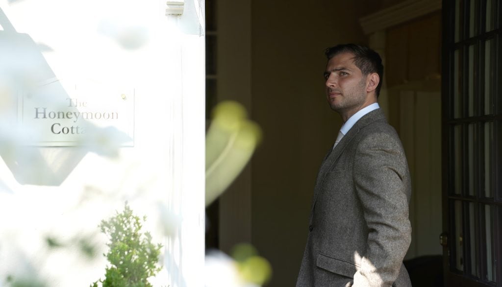 A man in a suit stands outside a building with a sign reading "The Honeymoon Cottage.