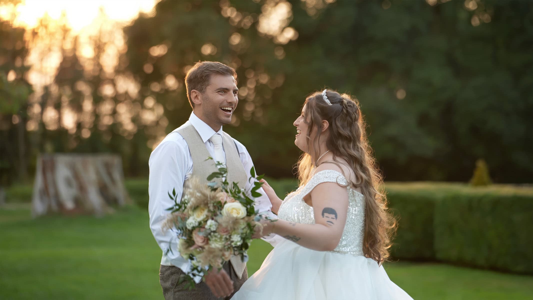 A couple stands in a garden at sunset, exchanging smiles. The woman holds a bouquet and wears a white dress, while the man is in a vest and tie.