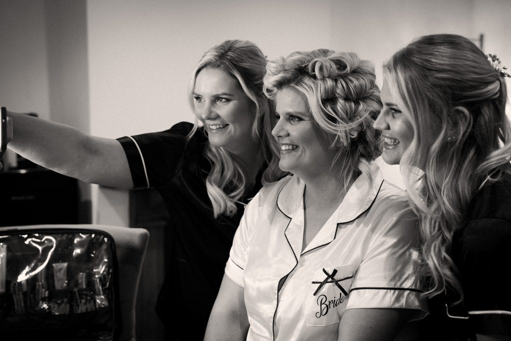 Three women in matching pajamas, one labeled "Bride," smile for a selfie indoors.