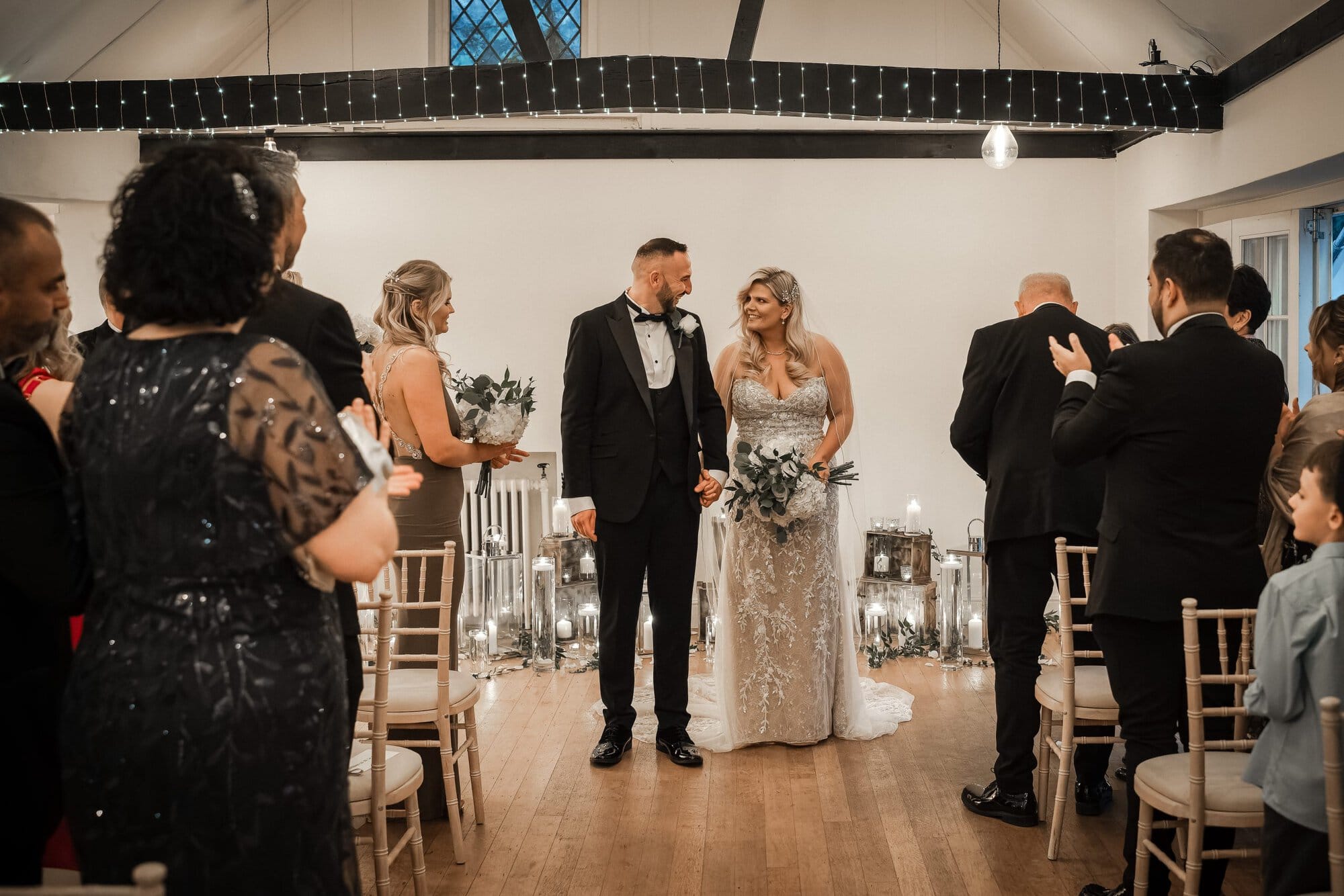A bride and groom stand at the altar holding hands, surrounded by formally dressed guests in a decorated indoor venue.