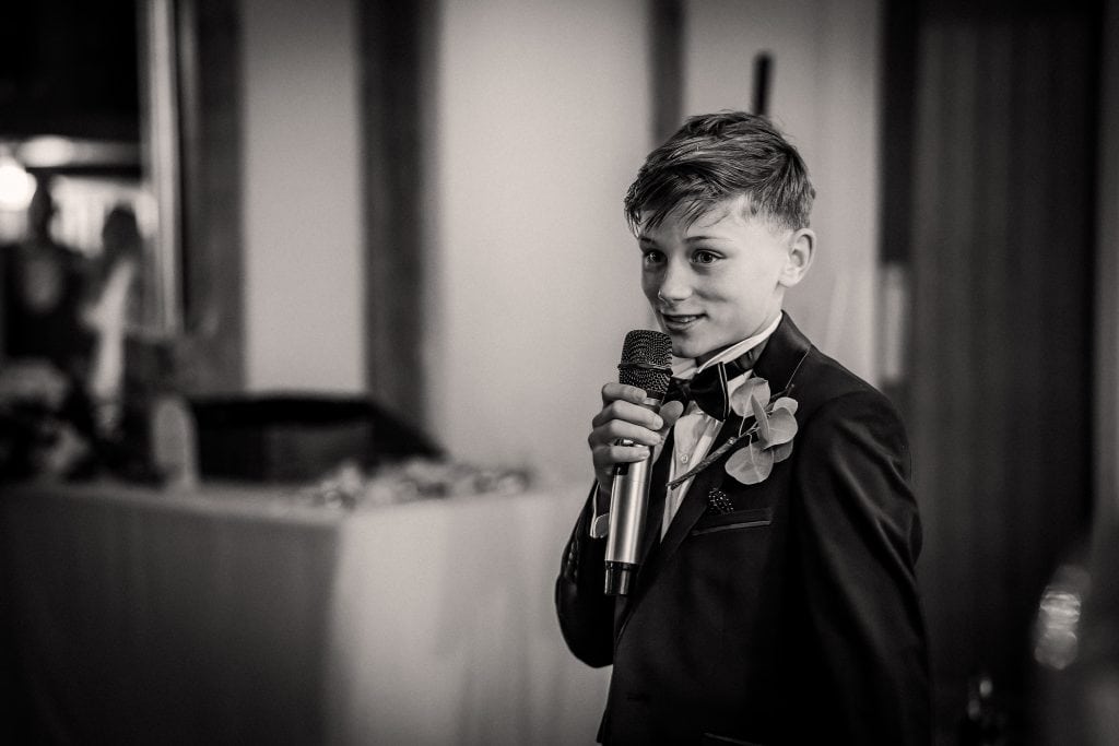 A young boy in a suit and bowtie holds a microphone, speaking indoors.