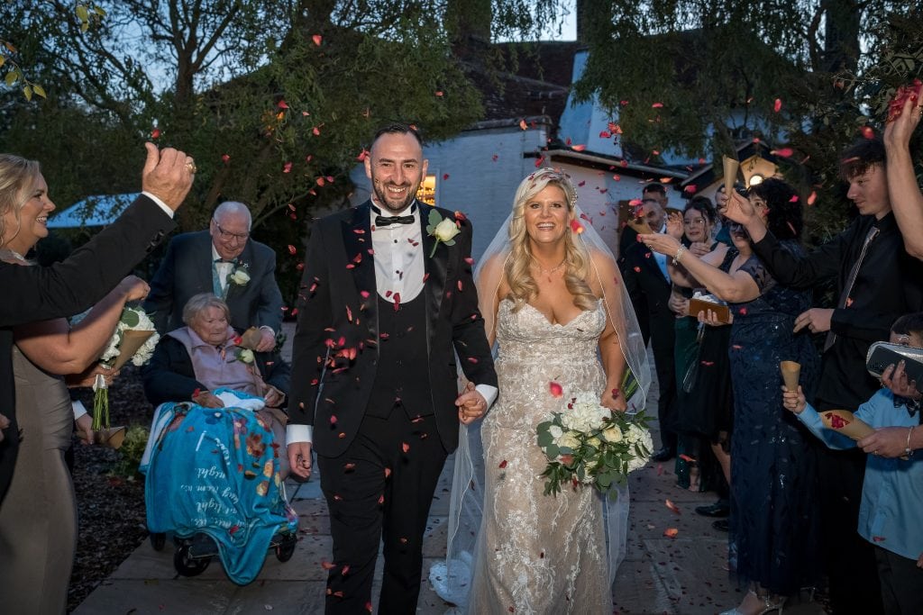 Bride and groom walk hand in hand with guests celebrating and tossing rose petals.