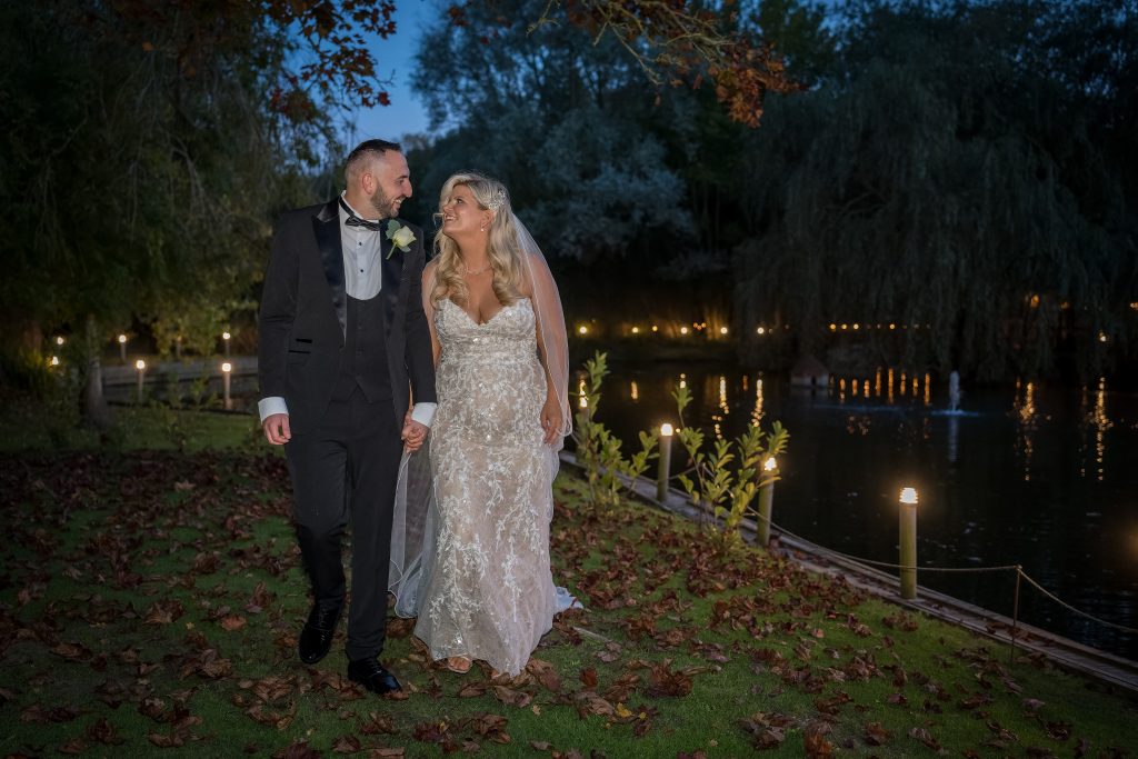 A bride and groom walk hand in hand on a grass path by a pond during the evening, surrounded by trees and soft lighting.