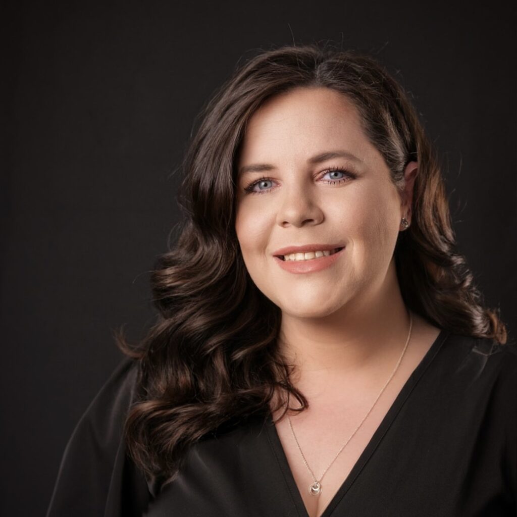 A person with long dark hair and a slight smile, wearing a black top and necklace, is posed against a dark background.