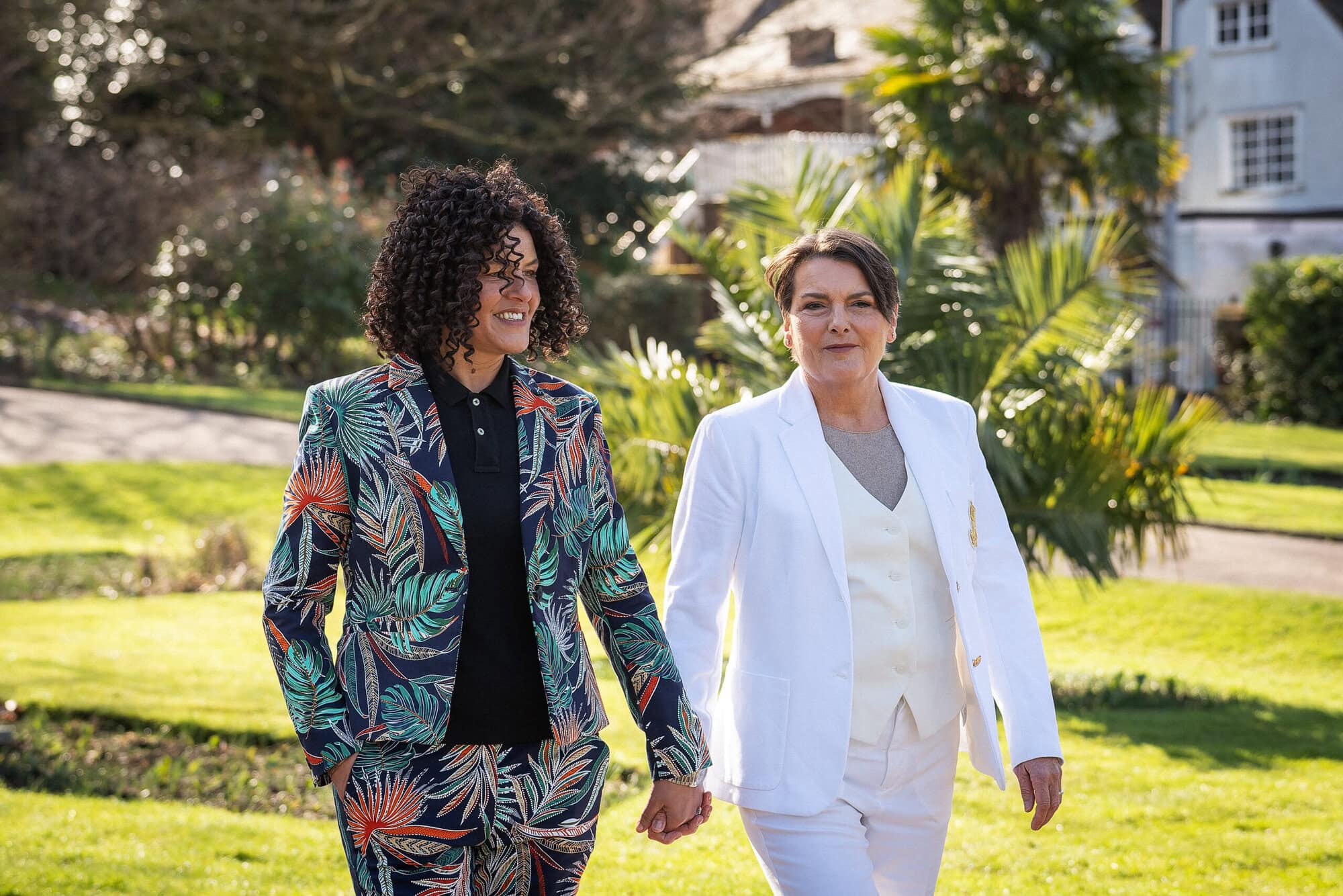 Two people walking outdoors holding hands, one in a colorful floral suit and the other in a white suit. They are in a garden-like setting with buildings in the background.
