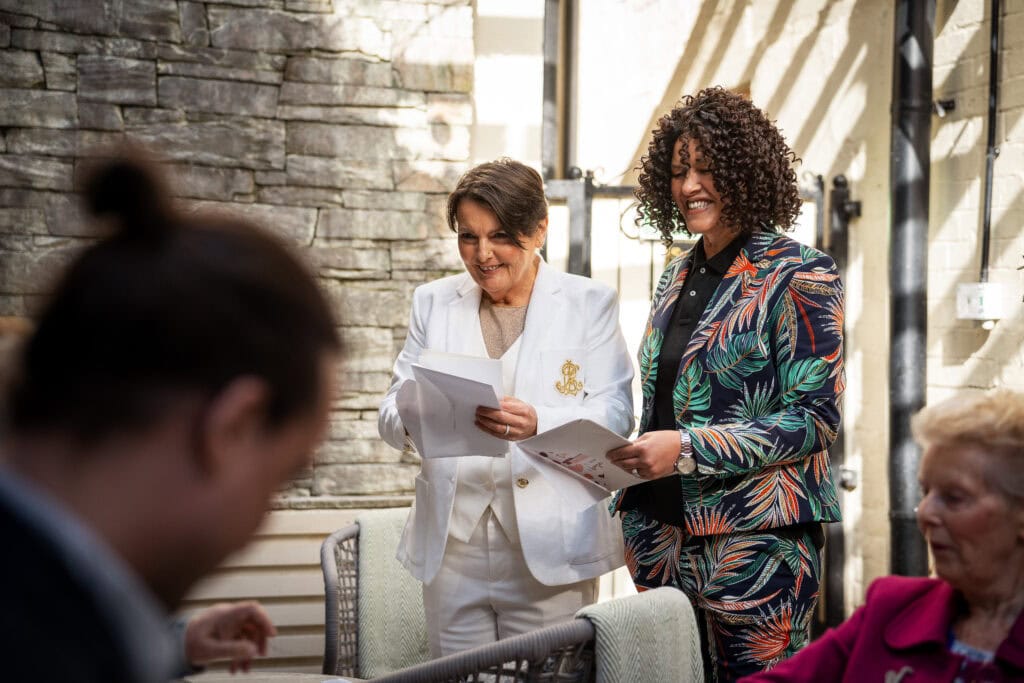 Two people smiling and holding papers, dressed in formal attire. One wears a white suit, the other a colorful one. They stand in front of a stone wall, with others seated nearby.