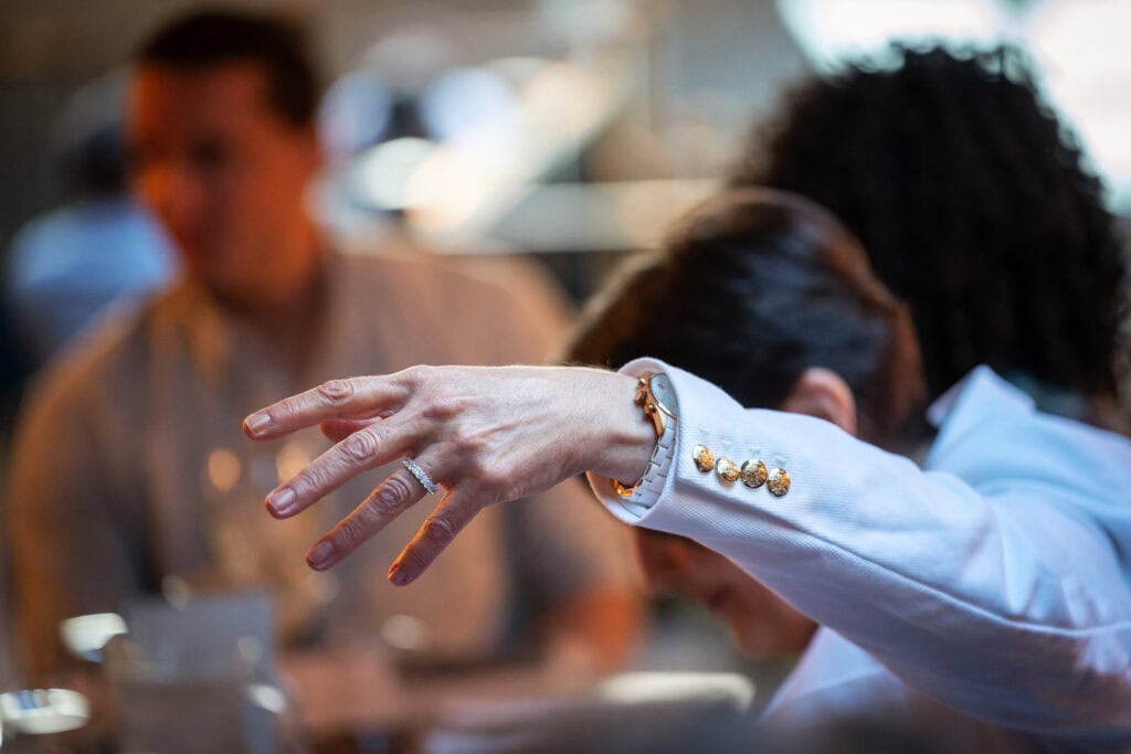 A person in a white suit, with gold buttons, extends their right arm outwards. Background shows blurred individuals in a dimly lit room.
