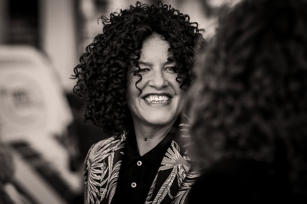 A person with curly hair smiling while standing outdoors, wearing a patterned jacket.