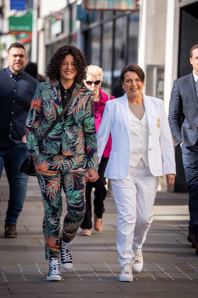 Two people walking hand in hand down a city street. One wears a colorful leaf-patterned suit, the other a white suit. They are smiling. Other pedestrians are visible in the background.