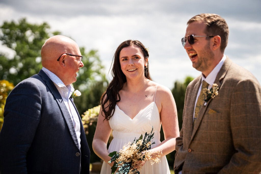 Three people stand outside at a wedding; a woman in a white dress holds a bouquet, flanked by two men in suits, one of whom is laughing.