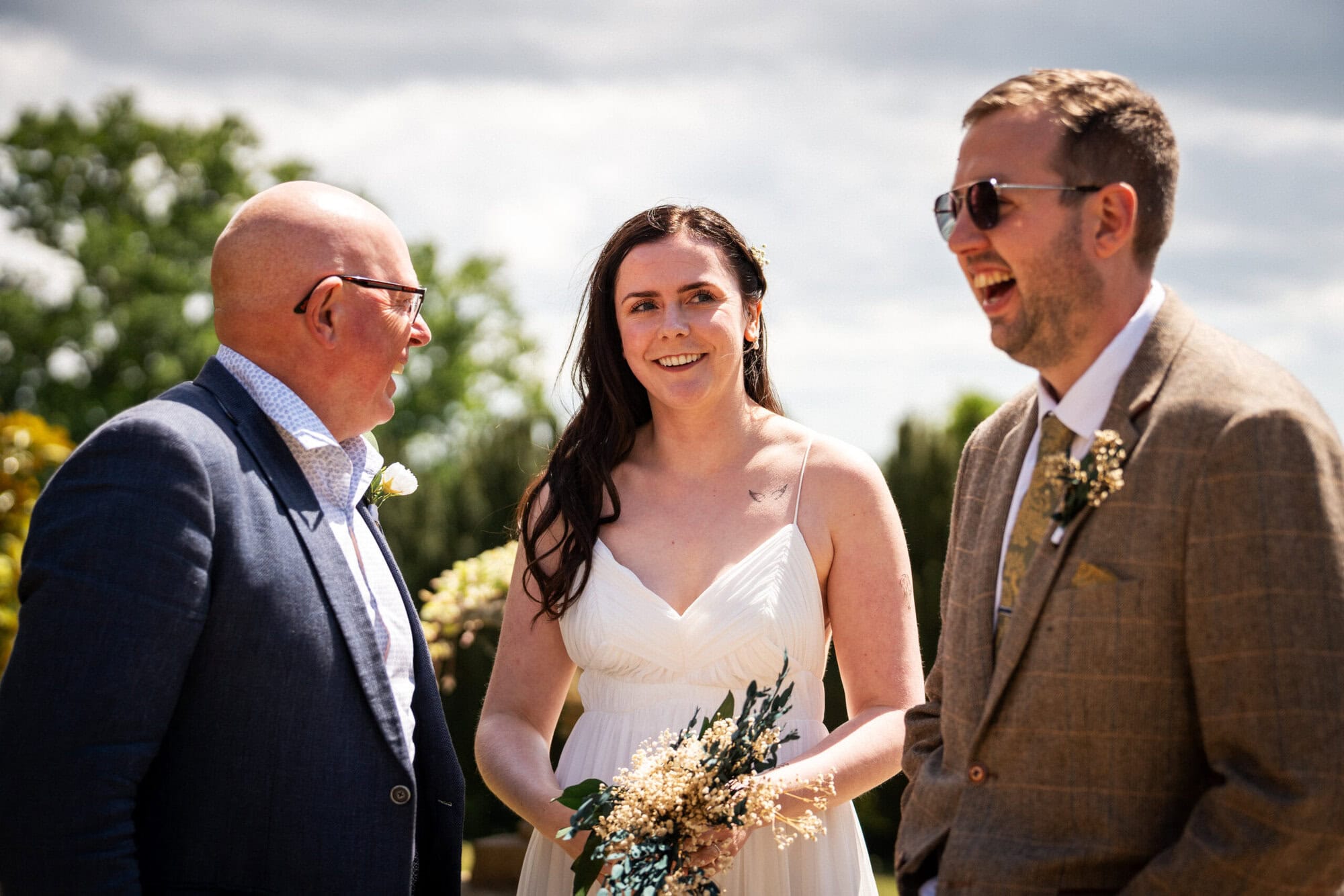 Three people stand outside at a wedding; a woman in a white dress holds a bouquet, flanked by two men in suits, one of whom is laughing.