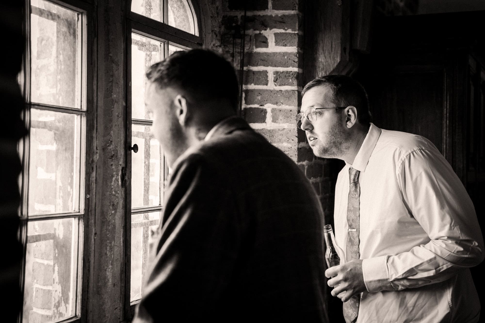 Two men stand indoors looking out a window; one is holding a bottle. They are dressed in collared shirts and jackets. The scene is in black and white.