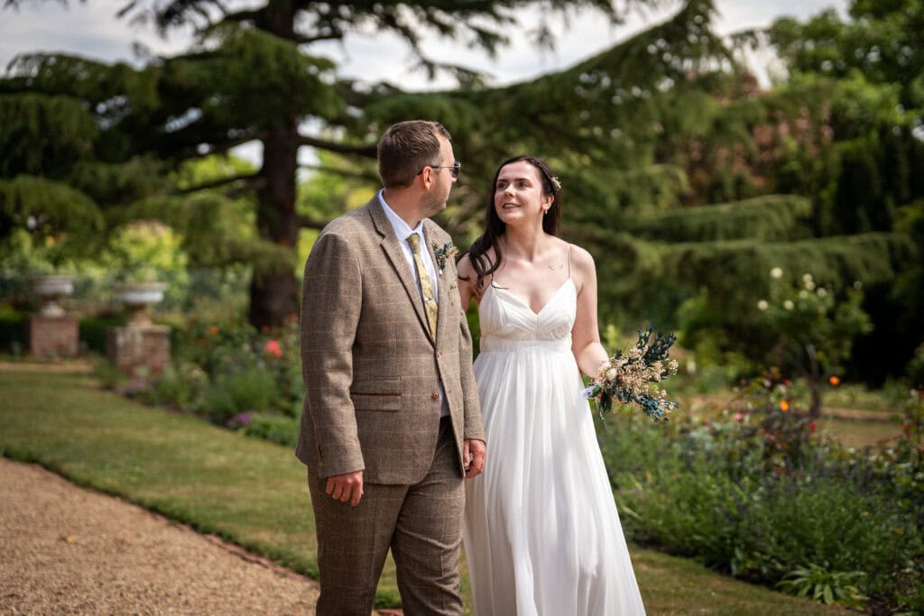 A bride in a white dress and a groom in a brown suit walk together outdoors along a garden path, surrounded by greenery and flowers.