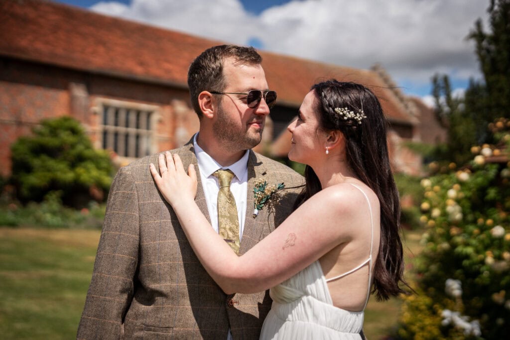 A bride in a white dress stands close to a groom in a checked suit and tie, outdoors on a sunny day, with greenery and a brick building in the background.