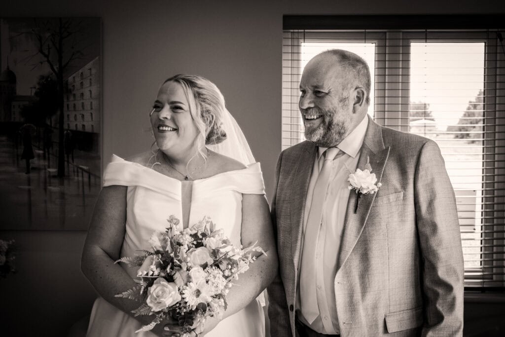 A bride in a white dress holding a bouquet stands beside a man in a light suit and boutonniere; both are smiling indoors by a window.