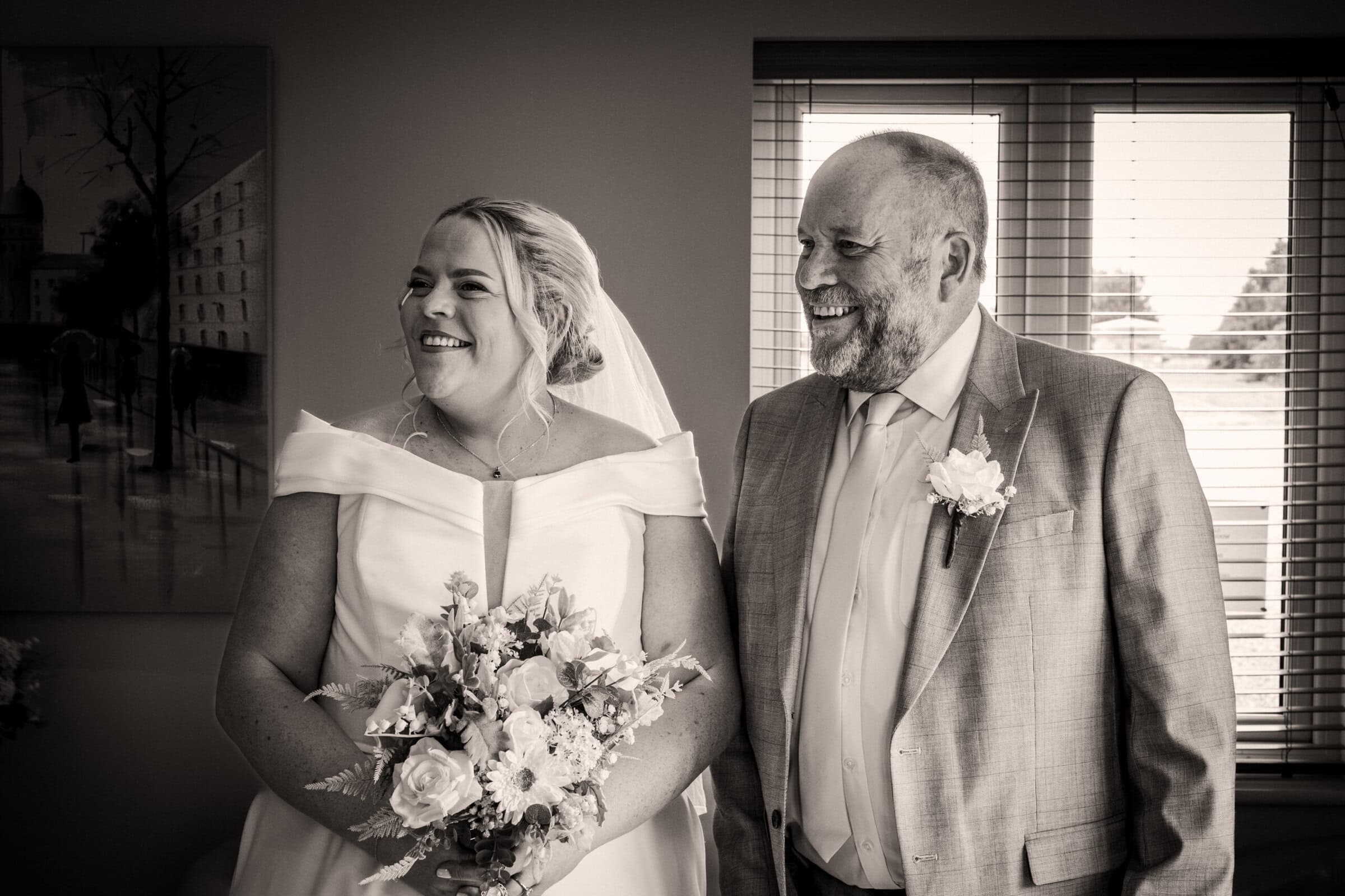 A bride in a white dress holding a bouquet stands beside a man in a light suit and boutonniere; both are smiling indoors by a window.