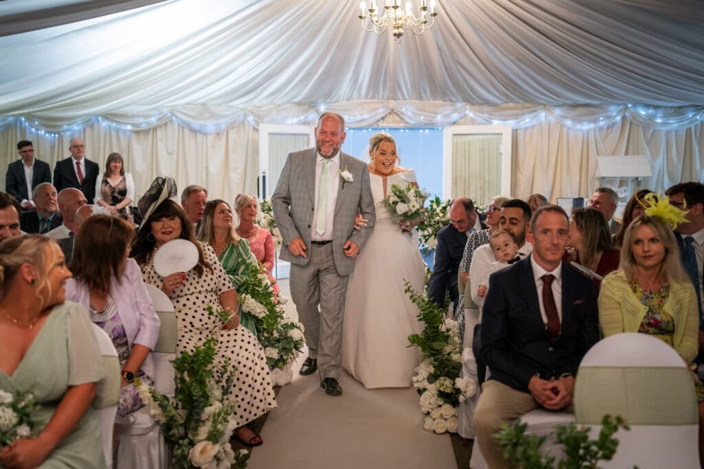 A bride in a white gown walks down the aisle on the arm of an older man in a gray suit, surrounded by seated guests in a decorated wedding tent.
