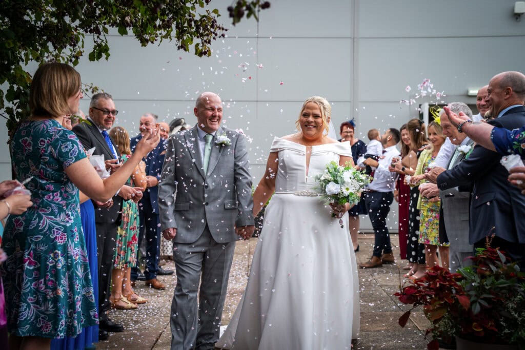A bride and groom walk hand in hand outside while guests throw confetti, celebrating their wedding.