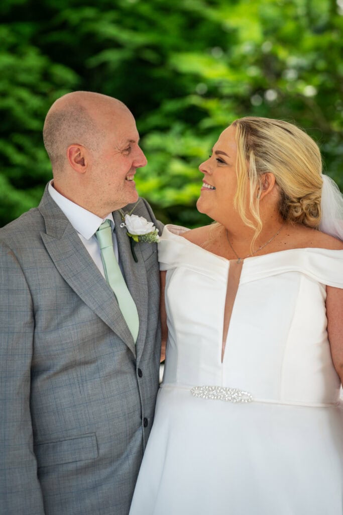 A bride in a white dress and a groom in a grey suit stand outdoors, facing each other and smiling, with greenery in the background.