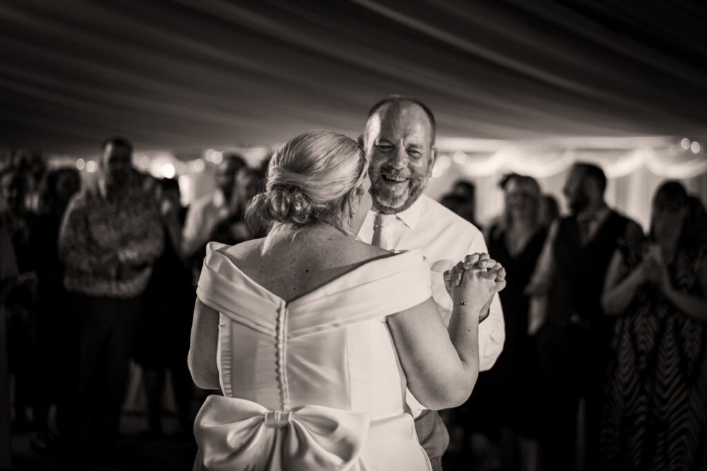 A couple dances together at an event, with the woman in a white dress and the man in a white shirt, surrounded by people in the background.