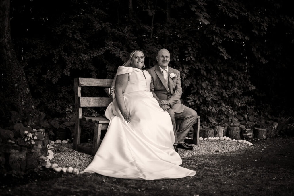 A bride and groom sit on a wooden bench outdoors, surrounded by trees and decorative stones, both smiling at the camera.