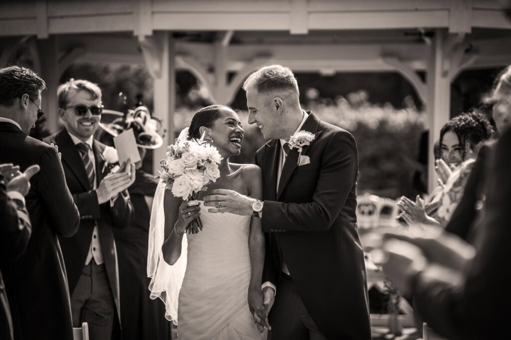 A bride and groom walk together, smiling and holding hands, as guests clap and celebrate during an outdoor wedding ceremony.