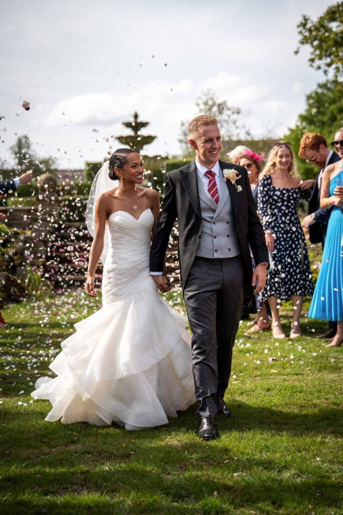 A bride and groom walk outdoors on grass, holding hands and smiling, as guests throw confetti around them at a wedding celebration.