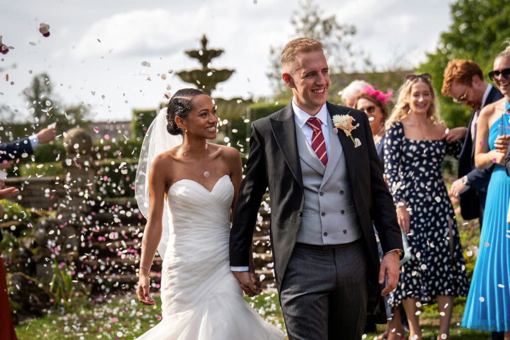 A bride and groom walk hand in hand outdoors as wedding guests throw flower petals, celebrating their marriage on a sunny day.