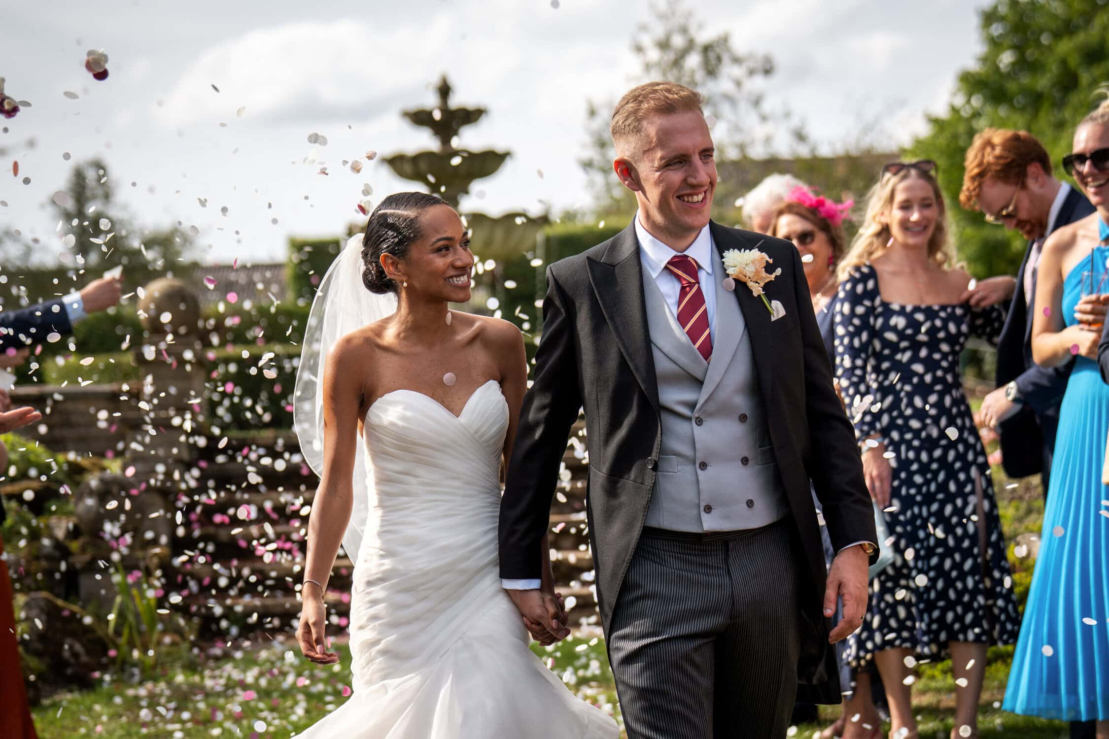 A bride and groom walk hand in hand outdoors as wedding guests throw flower petals, celebrating their marriage on a sunny day.