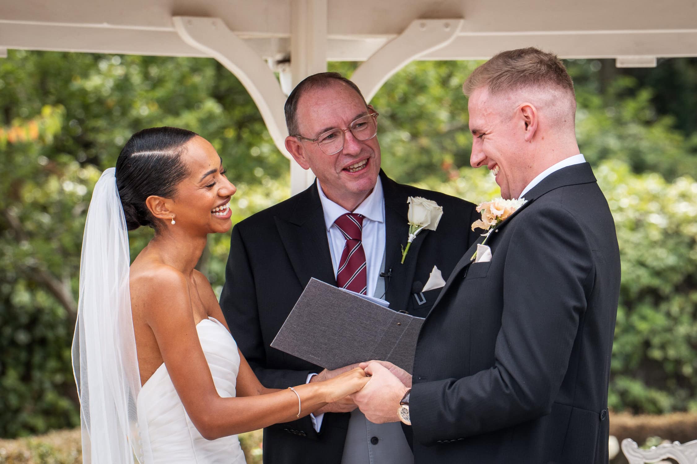 A bride and groom stand facing each other and holding hands, smiling as an officiant with a book stands between them during an outdoor wedding ceremony.