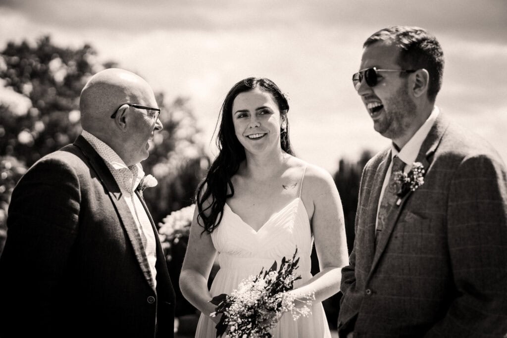Three adults stand together outdoors, dressed formally; the woman in a white dress holds flowers, while the two men, one in sunglasses, are smiling and engaged in conversation.