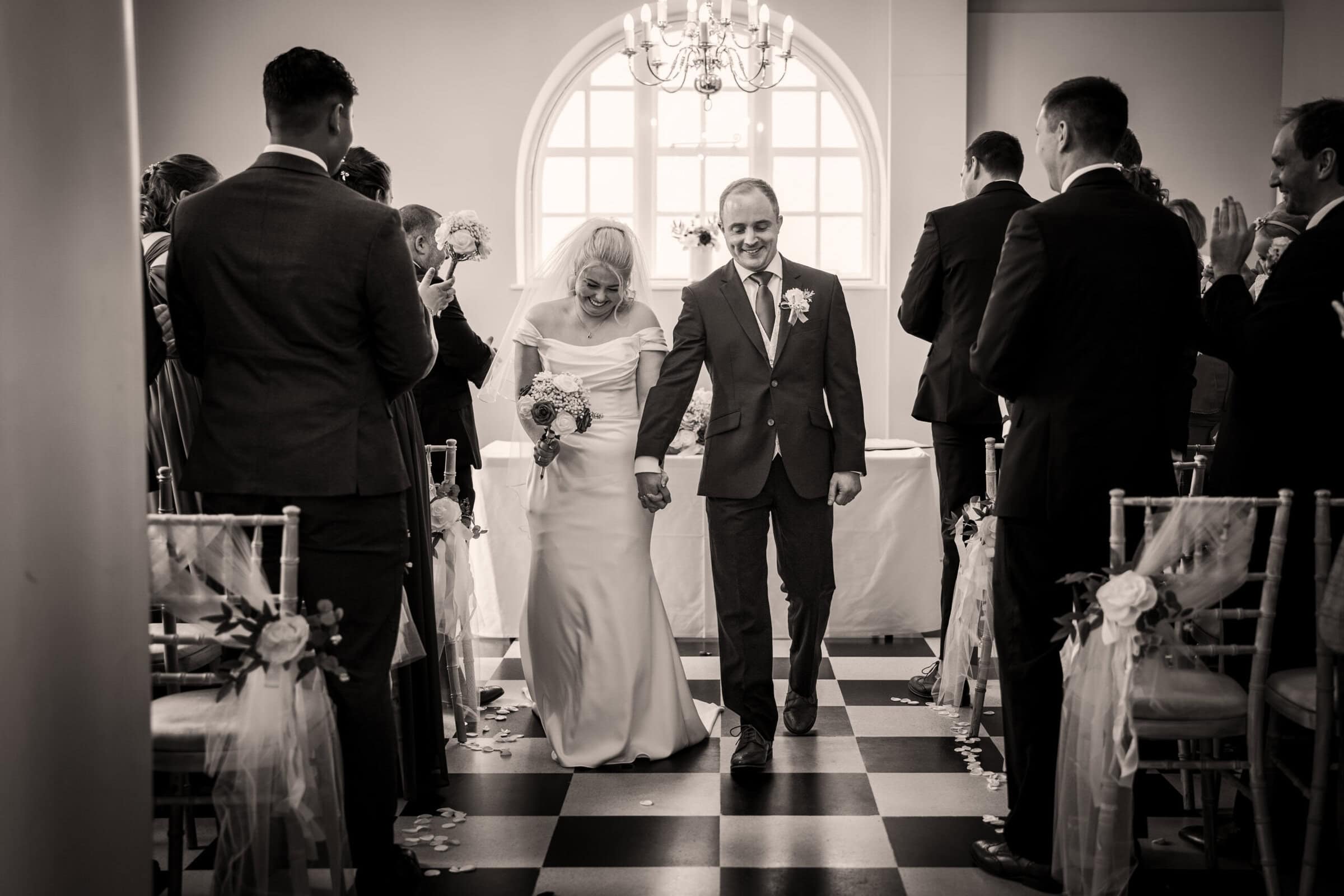 Samantha_Ollie_Wedding_Bury_St_Edmunds-420 A bride and groom walk down the aisle holding hands in a decorated indoor venue, while guests stand and applaud.