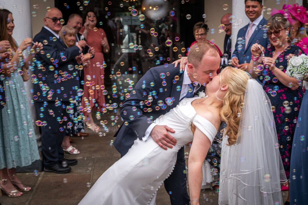 A bride and groom kiss while surrounded by wedding guests blowing bubbles outside, with the groom dipping the bride in her white dress and veil.