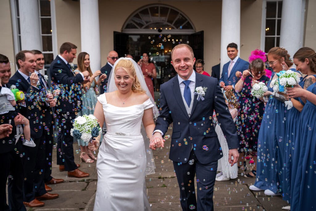 A bride and groom walk hand in hand outside, smiling, surrounded by guests blowing bubbles and holding bouquets.