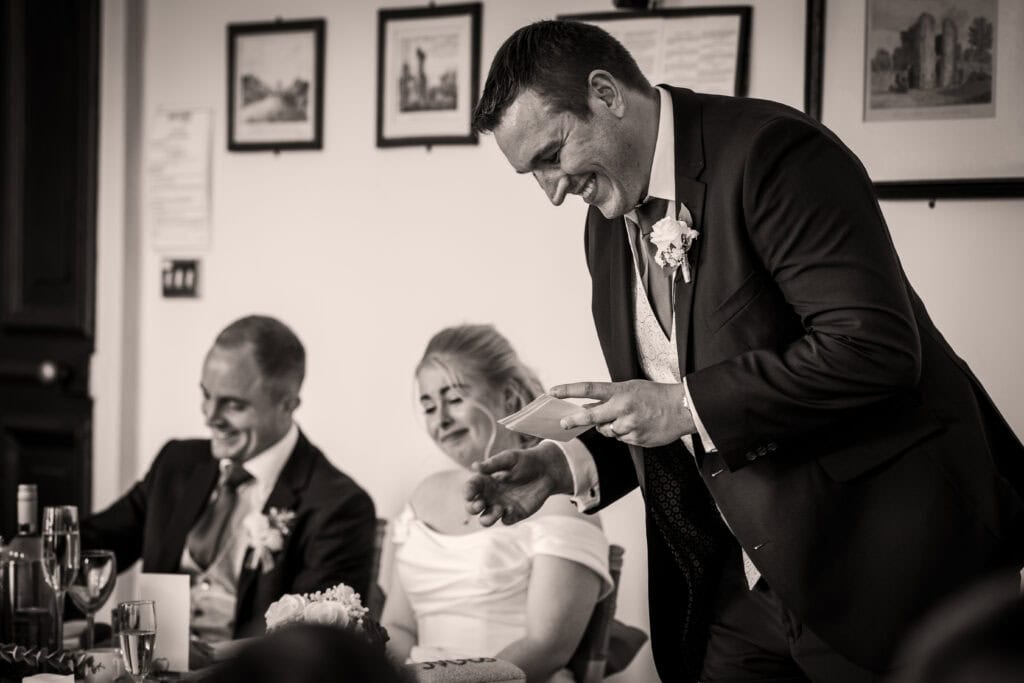 A man in a suit stands giving a speech while two seated people, also dressed formally, listen and smile at a wedding reception.
