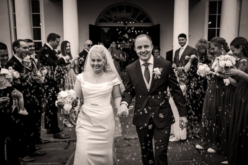 A bride and groom walk outside a building, smiling and holding hands, surrounded by guests blowing bubbles.