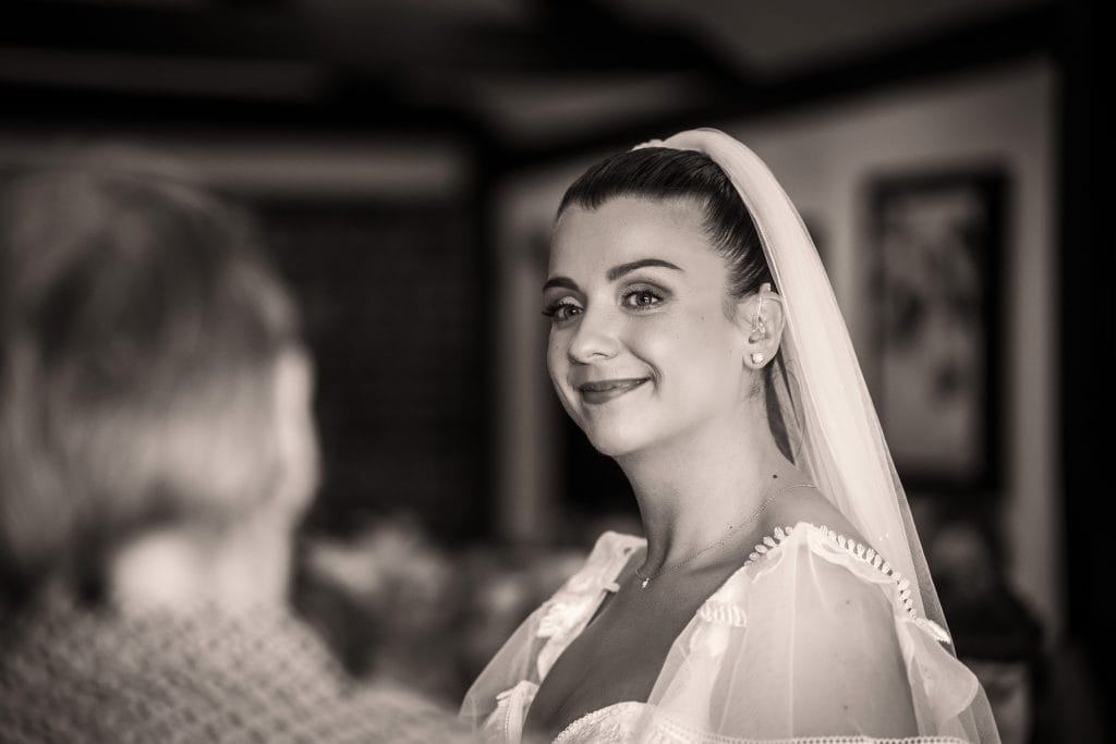 A woman in a wedding dress and veil smiles, looking toward the camera, with another person in the foreground.